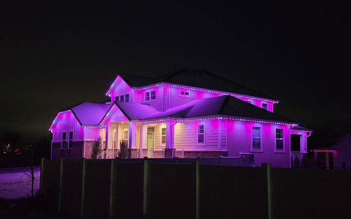 Two-story home glowing with vibrant pink and purple permanent LED Christmas lights along the rooflines and eaves at night, installed by Legacy Ridge Exteriors.